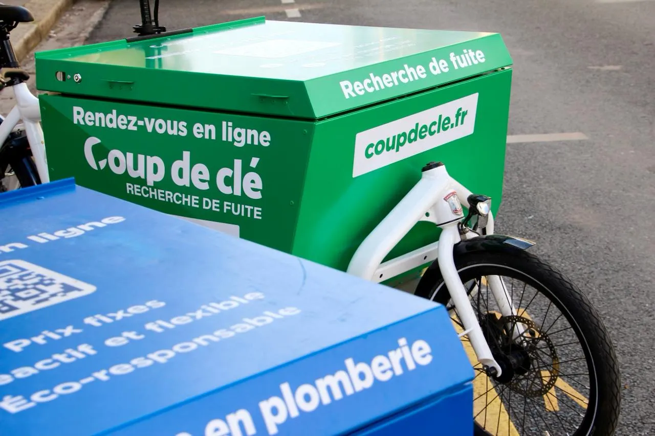 Vélo cargo blanc avec une grande caisse verte affichant « Rendez-vous en ligne Coup de clé Recherche de fuite » et une adresse web coupdeclé.fr, stationné sur une chaussée.