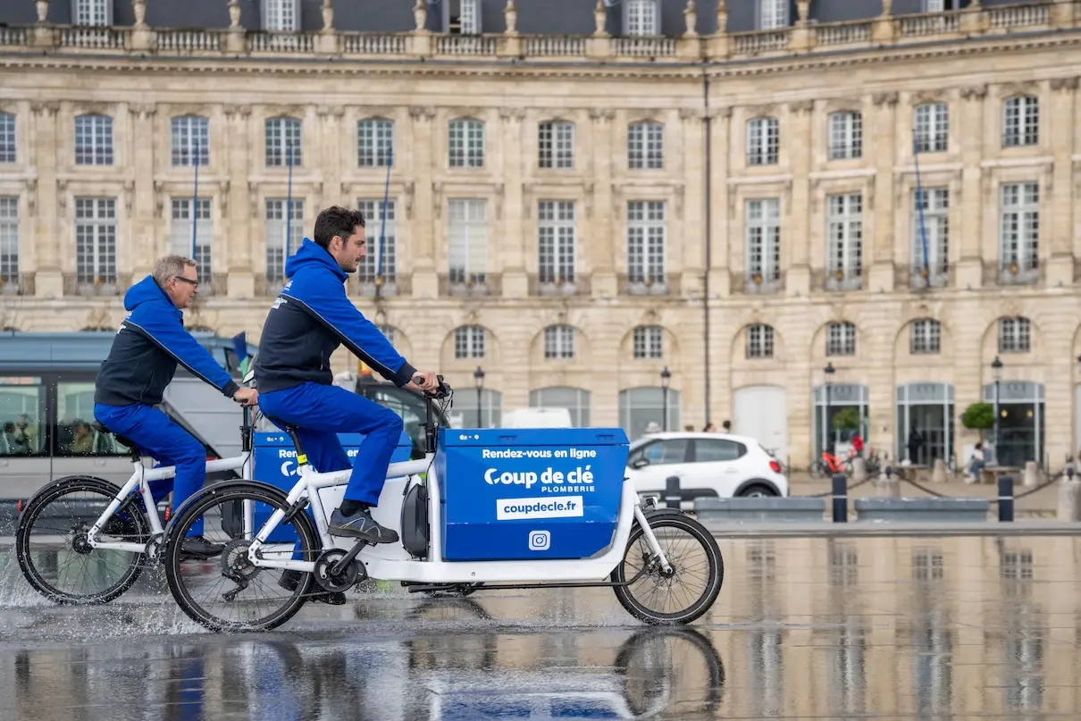 Deux hommes en tenue bleue à vélo cargo blanc avec publicité pour Coup de clé dans une rue de Rennes.