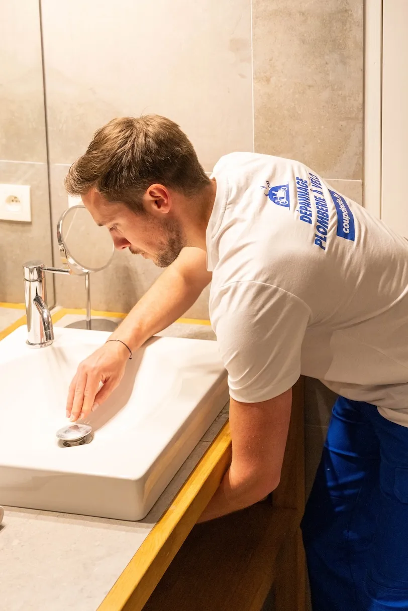 Un plombier en chemise blanche et pantalon bleu débouche un lavabo blanc dans une salle de bain avec mur carrelé beige.