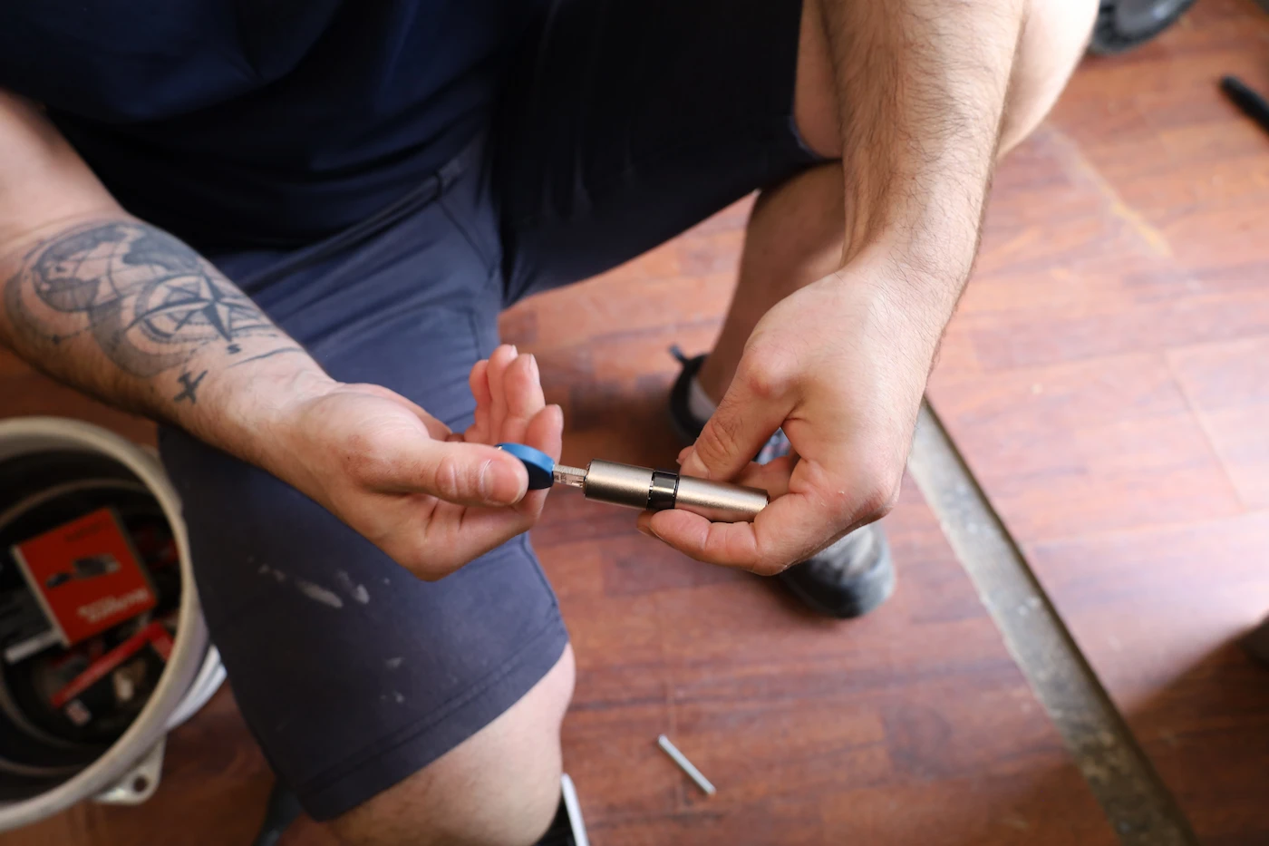 Un homme tatoué tenant un cylindre de serrure et une clé bleue dans ses mains, assis sur un sol en bois.