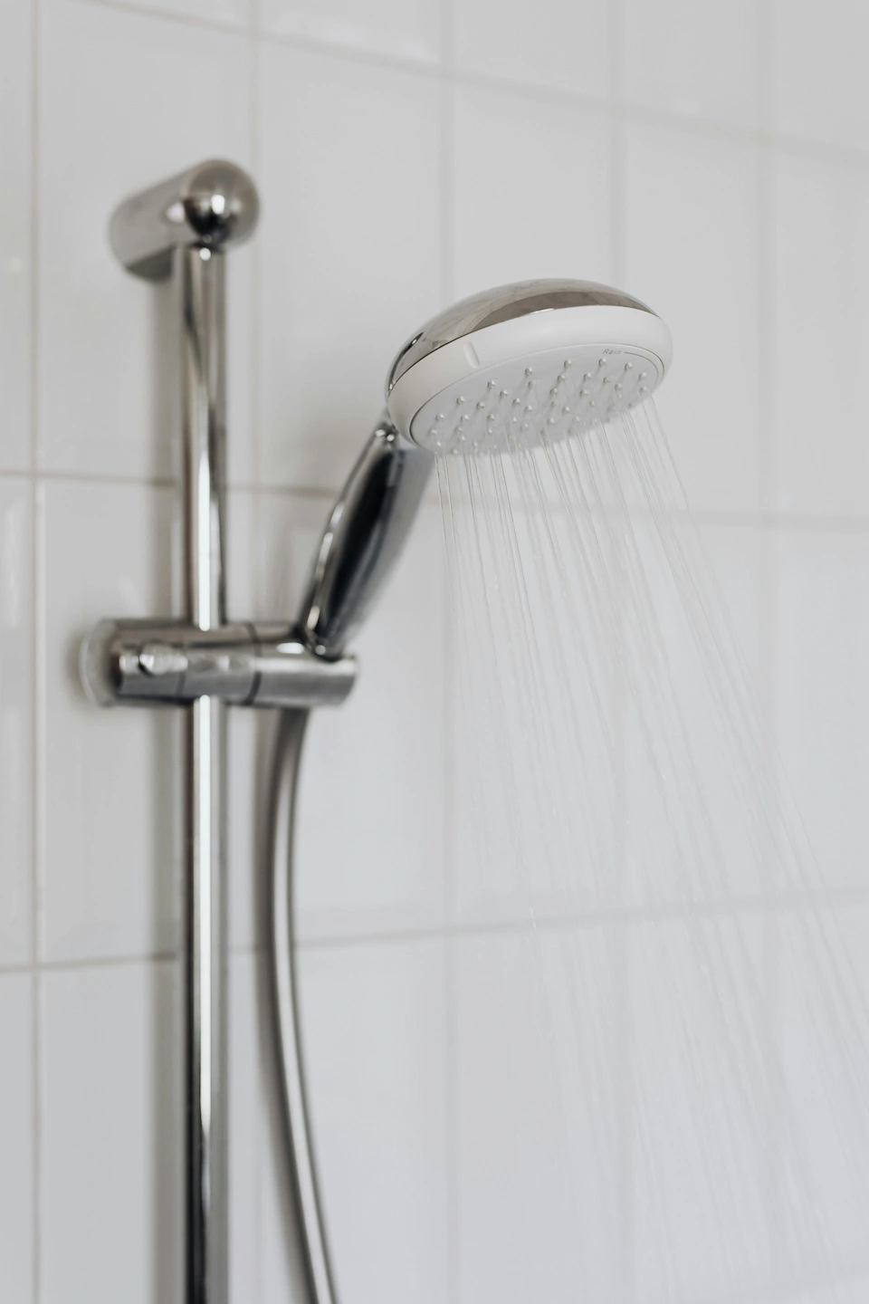 Douchette de salle de bain en acier inoxydable avec de l'eau qui coule, fixée sur un mur carrelé blanc.