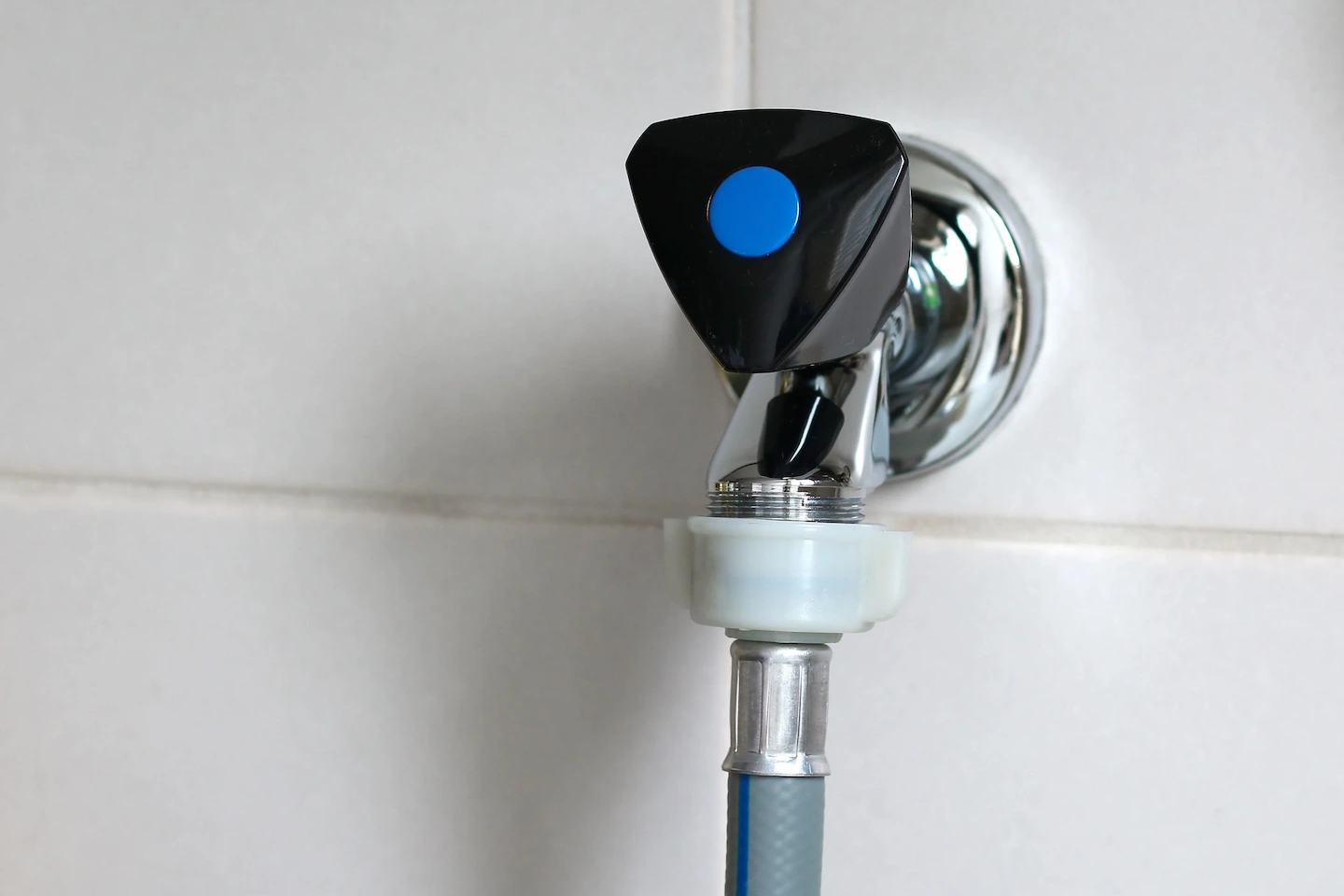 Robinet mural en métal chromé avec un bouton de commande noir et bleu raccordé à un tuyau gris.