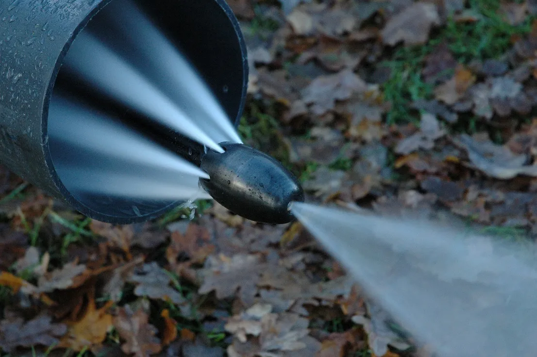 Jet d'eau à haute pression émanant d'une buse pour débouchage hydraulique, avec un sol couvert de feuilles en arrière-plan.