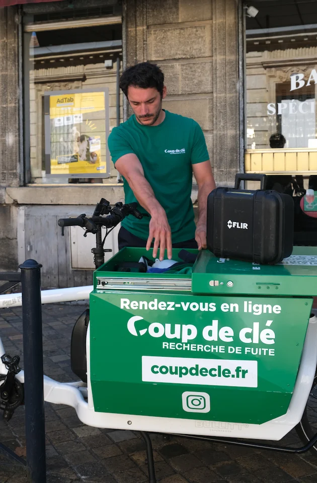 Homme portant un t-shirt vert avec le logo Coup de clé, travaillant sur un vélo cargo publicitaire pour la recherche de fuite, avec un boîtier FLIR posé dessus.