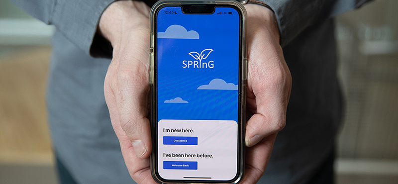 A closeup of a man holding a smartphone with the final Spring app's home screen displayed. The Spring logo is visible on a field of blue with light blue clouds. On the bottom third of the screen is an area with a white background. Two sections of text are visible: The first says "I'm new here" and a button underneath says "Get Started." The second says "I've been here before" and a button underneath says "Welcome Back."