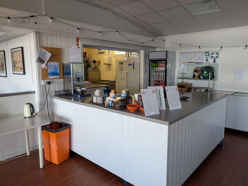 Indoor cafe counter with a menu, cups, a kettle, and a refrigerator stocked with drinks in a clean, organised space.