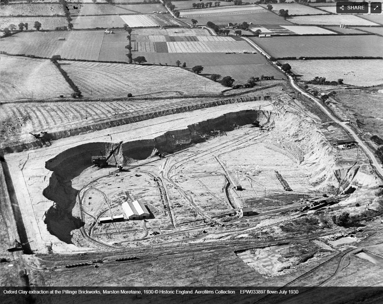 Aerial black and white photo of an open clay extraction site surrounded by farmland with visible excavation equipment and tracks.