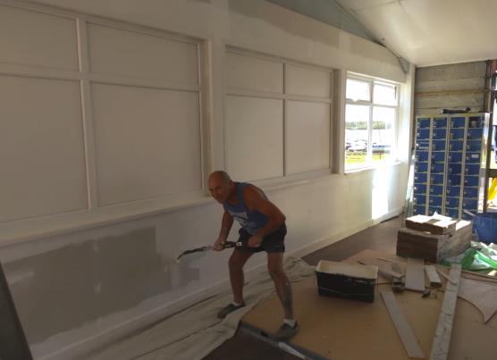 Man painting the lower part of an indoor wall in a workshop or storage area with tools and materials on the floor.