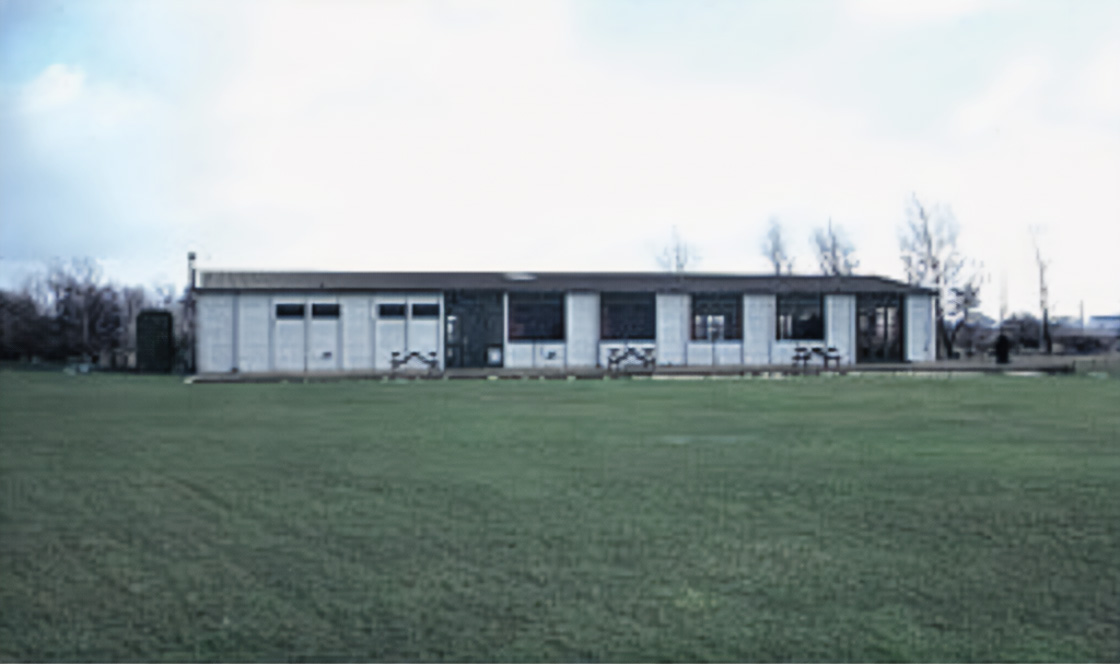 Main clubhouse. Single-storey building with large windows and picnic tables on a wide green lawn under a cloudy sky.