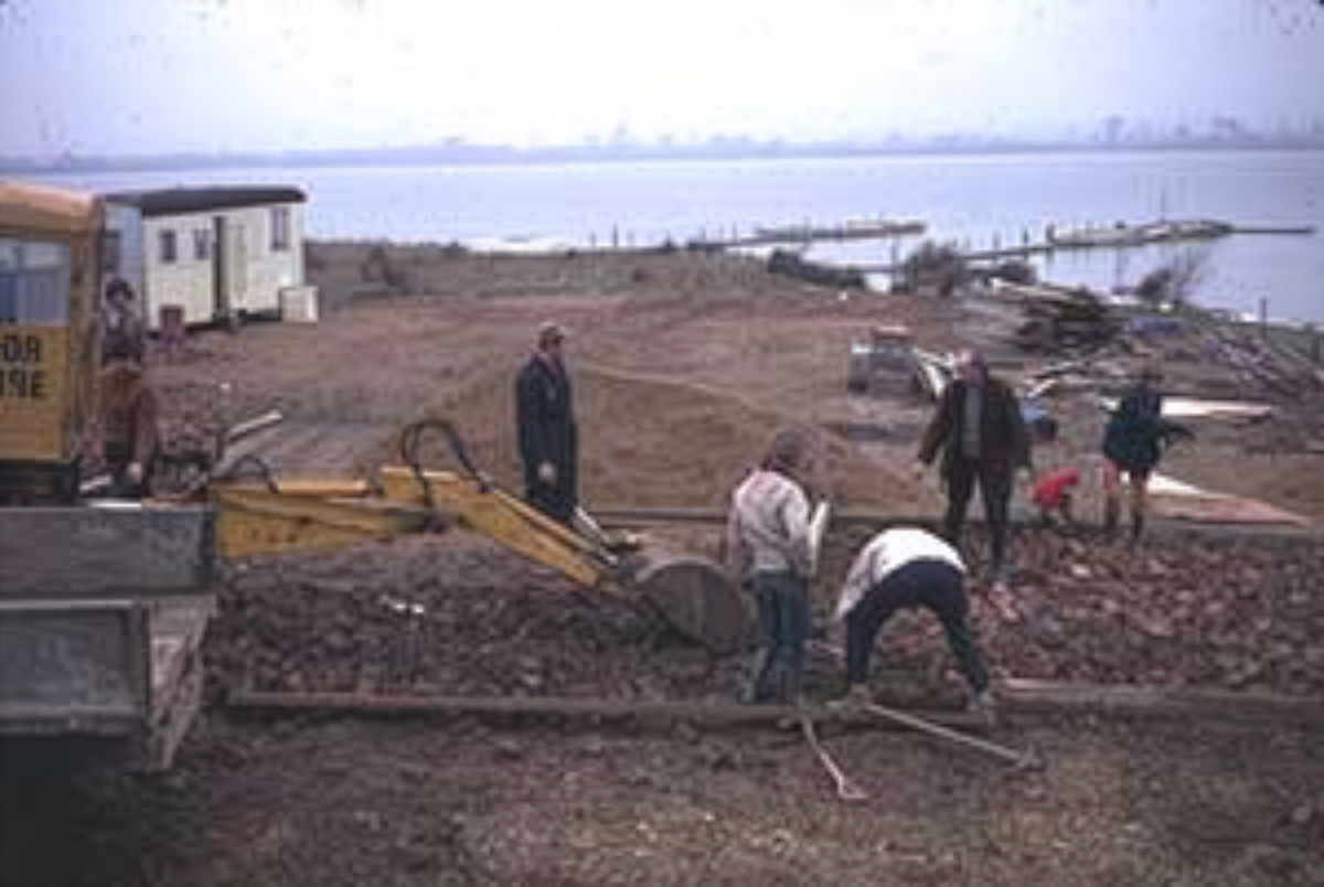 Construction workers using a small excavator and tools to work on a gravel surface near water with jettys in the background.