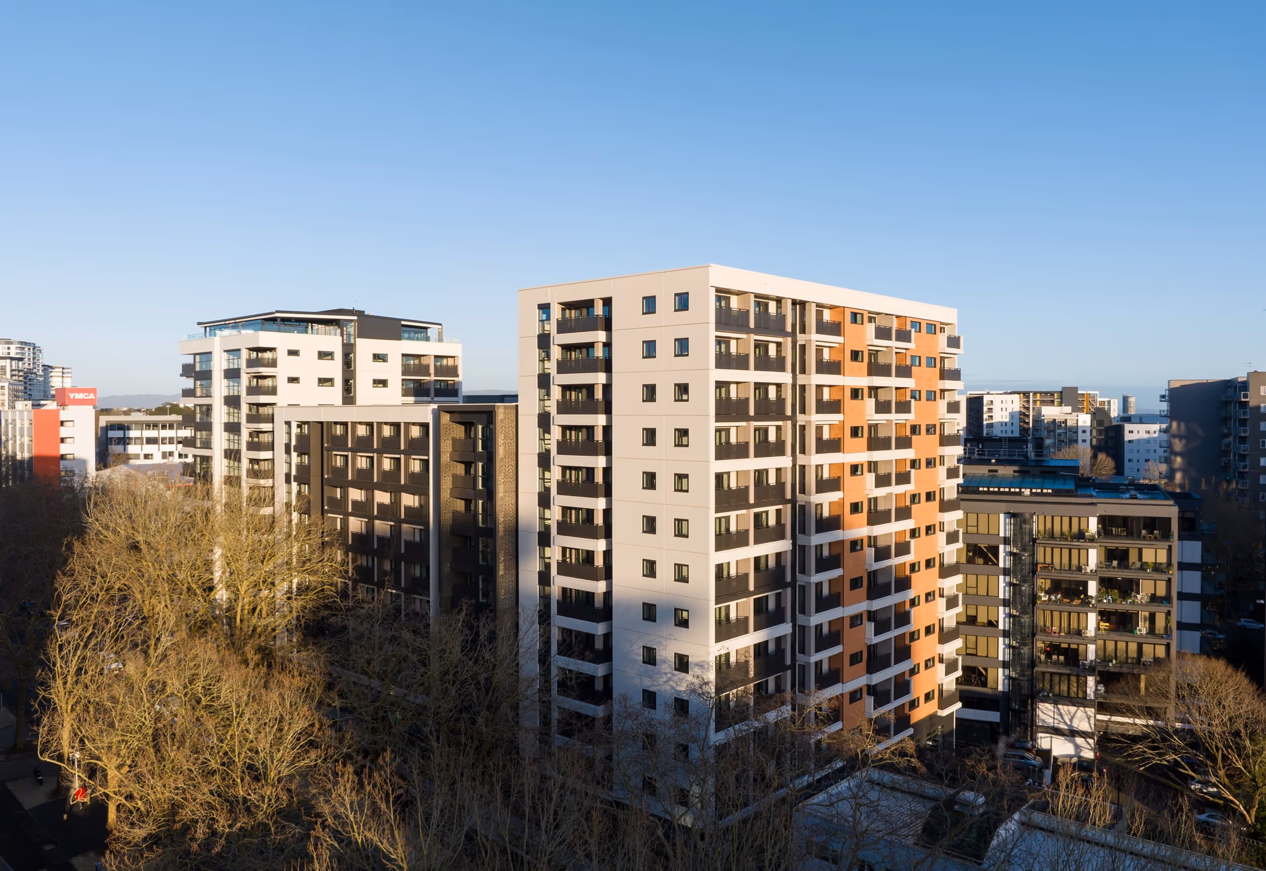 Cityscape showing several modern high-rise apartment buildings with trees in the foreground under a clear blue sky.