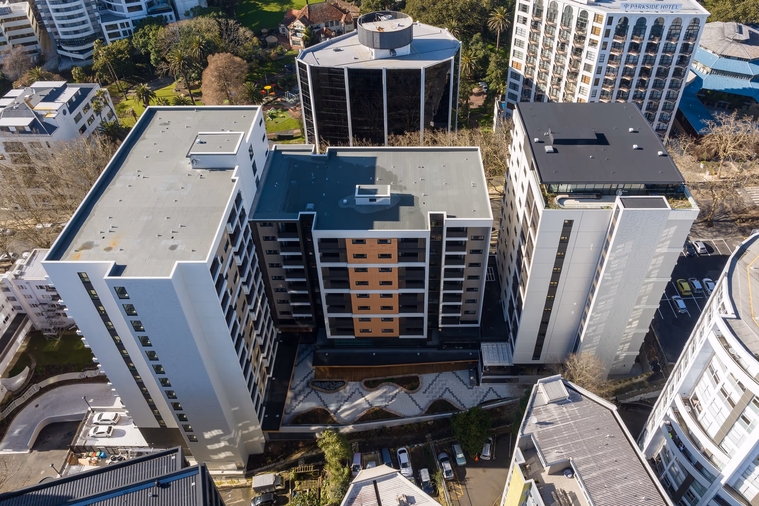 Aerial view of a modern urban area with three high-rise buildings. The buildings feature rectangular designs in grey and brown tones, surrounded by trees and roads. Nearby structures and parked cars are visible.