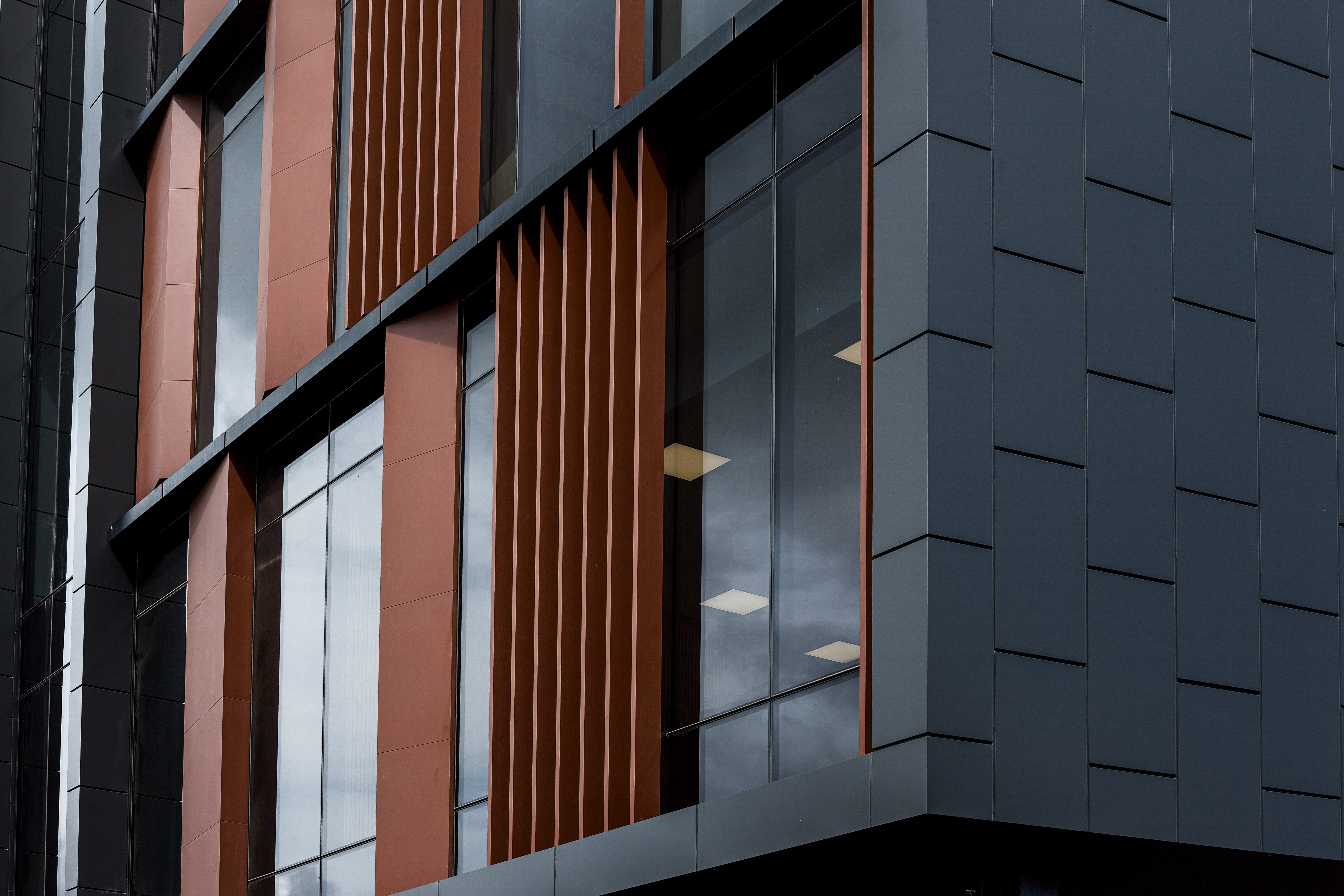 Close-up of Union Square Building E in Hamilton, New Zealand, showing the detailed façade design with terracotta-toned cladding, vertical fins, and dark metal panelling. The composition highlights the depth, texture, and precision of the envelope detailing.