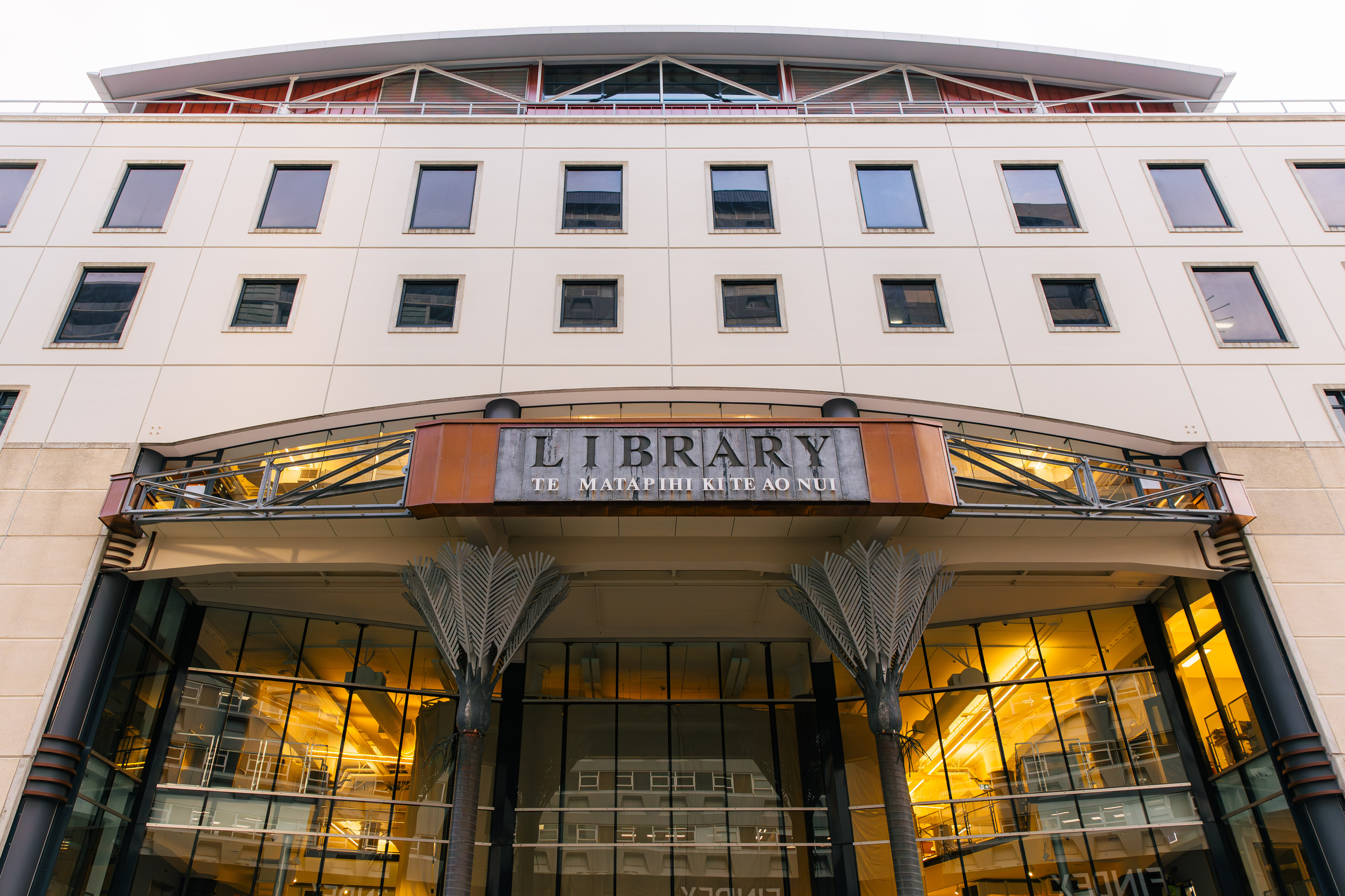 Contemporary library building featuring a glazed entrance, palm-shaped metal columns, and a sign reading “Library – Te Matapihi Ki Te Ao Nui.”