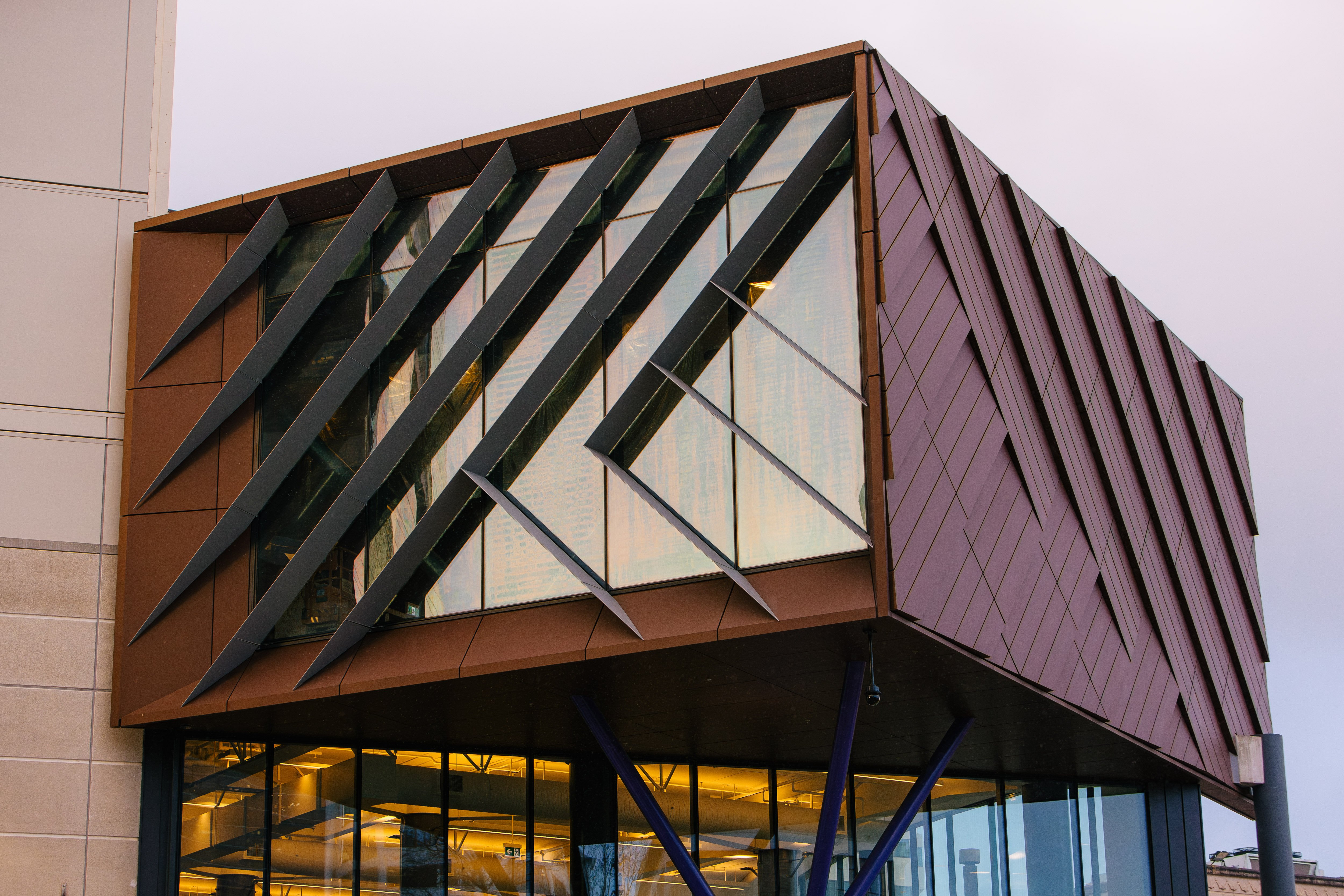 Contemporary building detail showing an angular glass façade framed by diagonal metal fins and copper-toned cladding, supported by slender steel columns below.