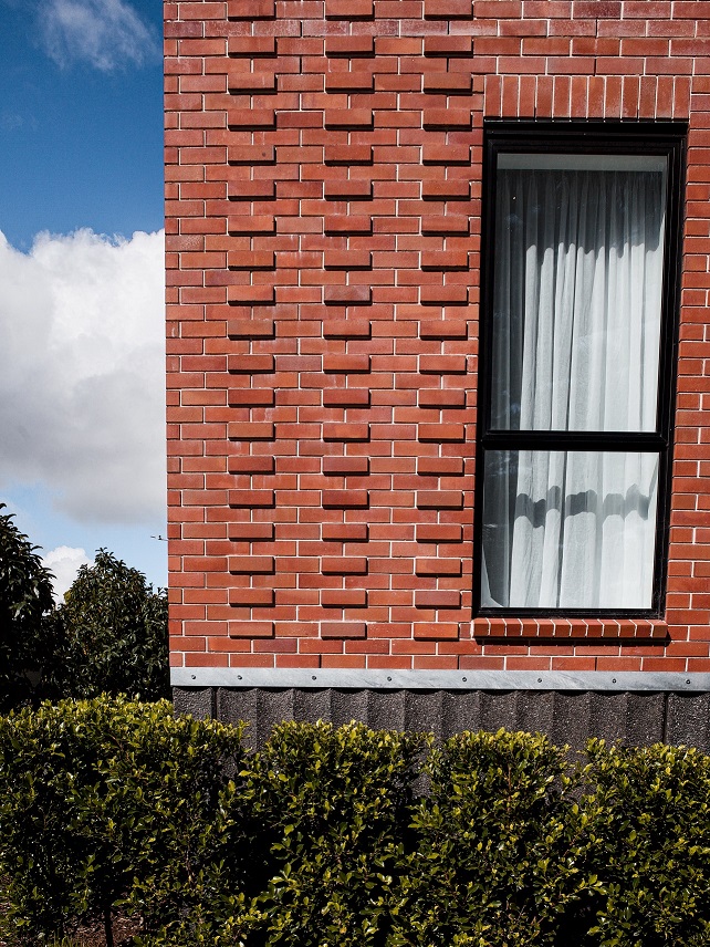 Corner of a red brick building with textured masonry, a tall window, and green bushes.