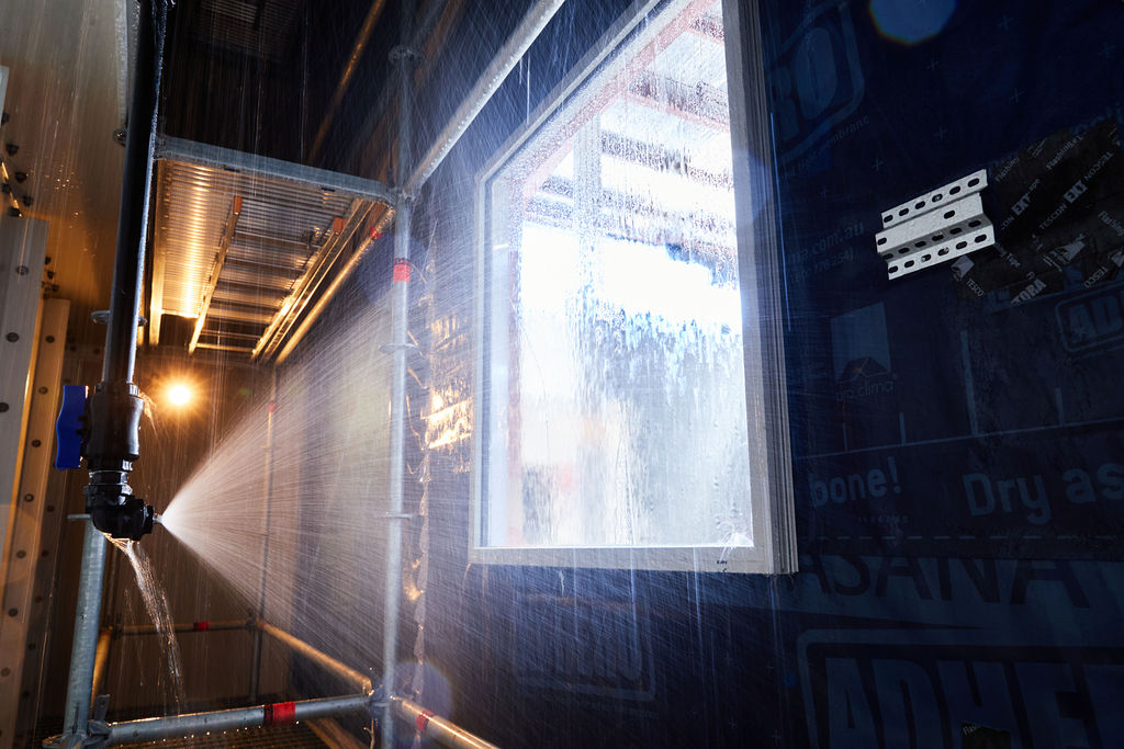 Interior view of a high-pressure water spray test being conducted on a window installed in a wall with 'pro clima' weather-resistive barrier during construction