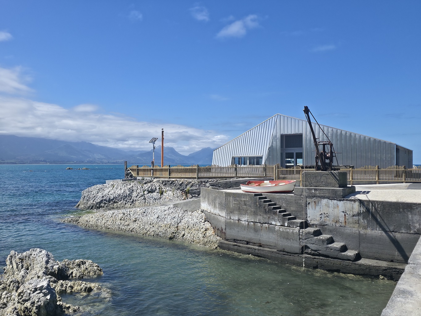 A wide view of the Wakatu Quay hospitality precinct in Kaikōura, where modern boat-hull architecture sits alongside historic wharf elements like a vintage crane.