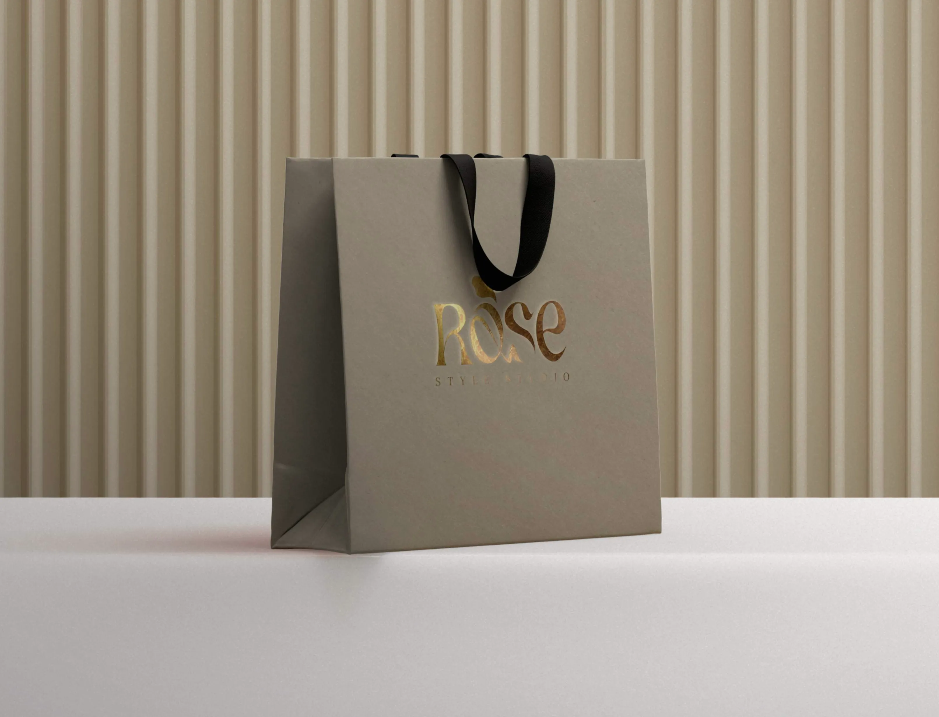 Gray shopping bag with black handles and gold embossed text reading 'ROSE STYLE STUDIO' on a white surface with a beige ribbed background.