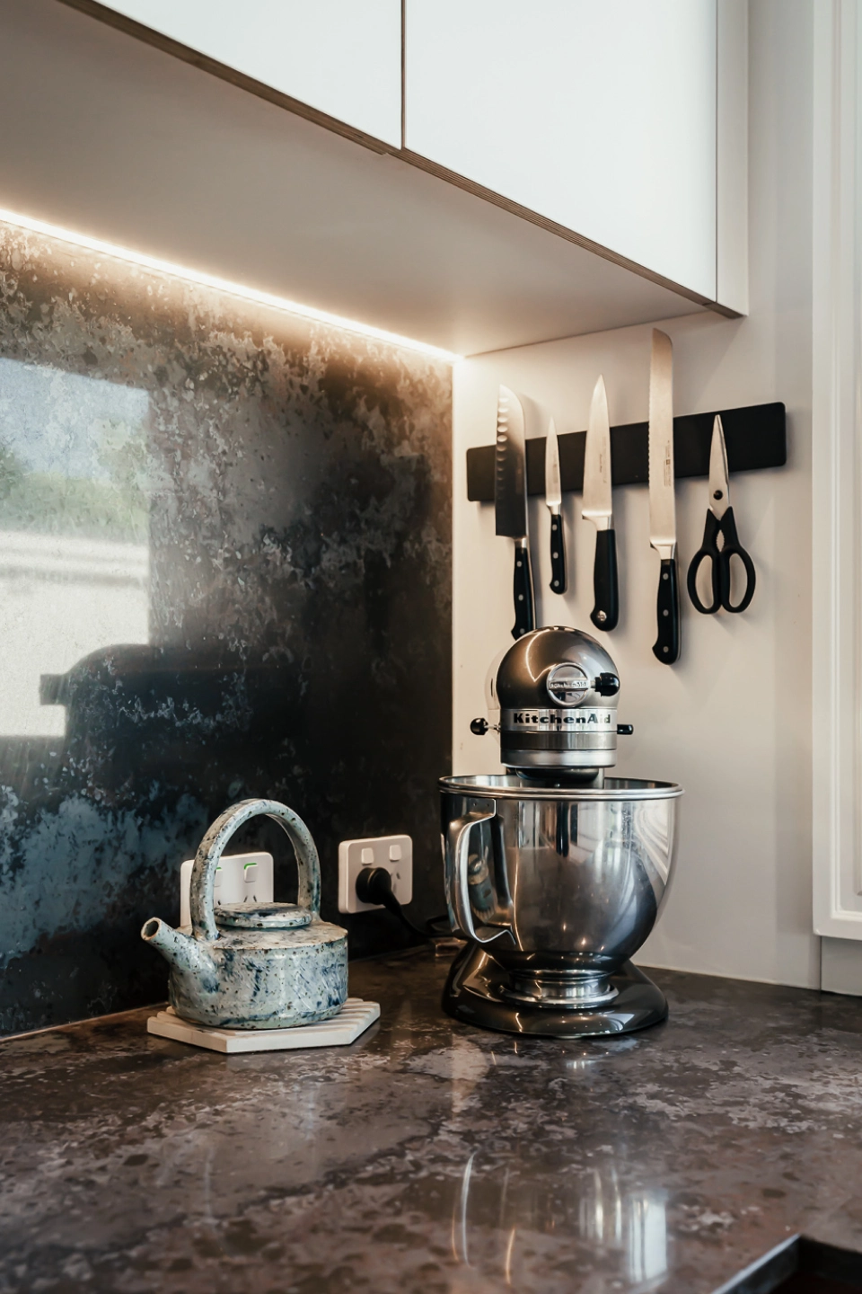 A modern dark bench displaying a stand mixer, teapot and a knife rack.