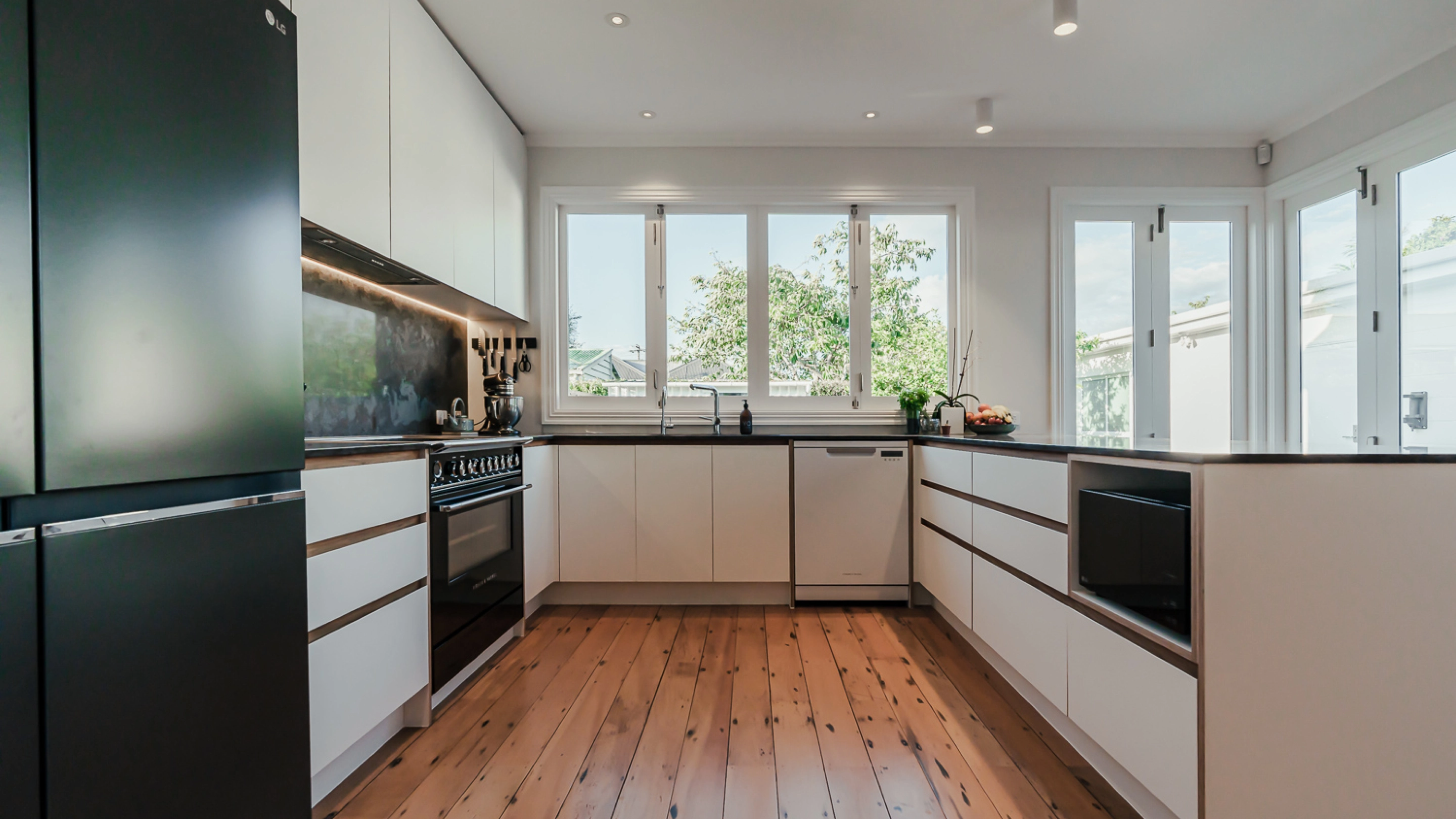 A bright, open kitchen with large windows, minimalist white cabinets, and black countertops.