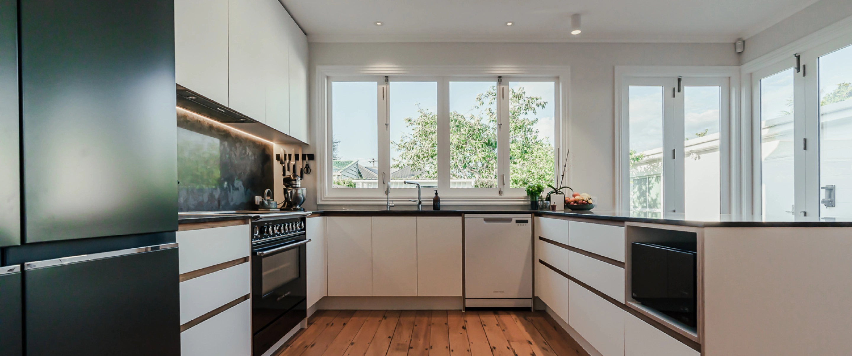 A bright, open kitchen with large windows, minimalist white cabinets, and black countertops.