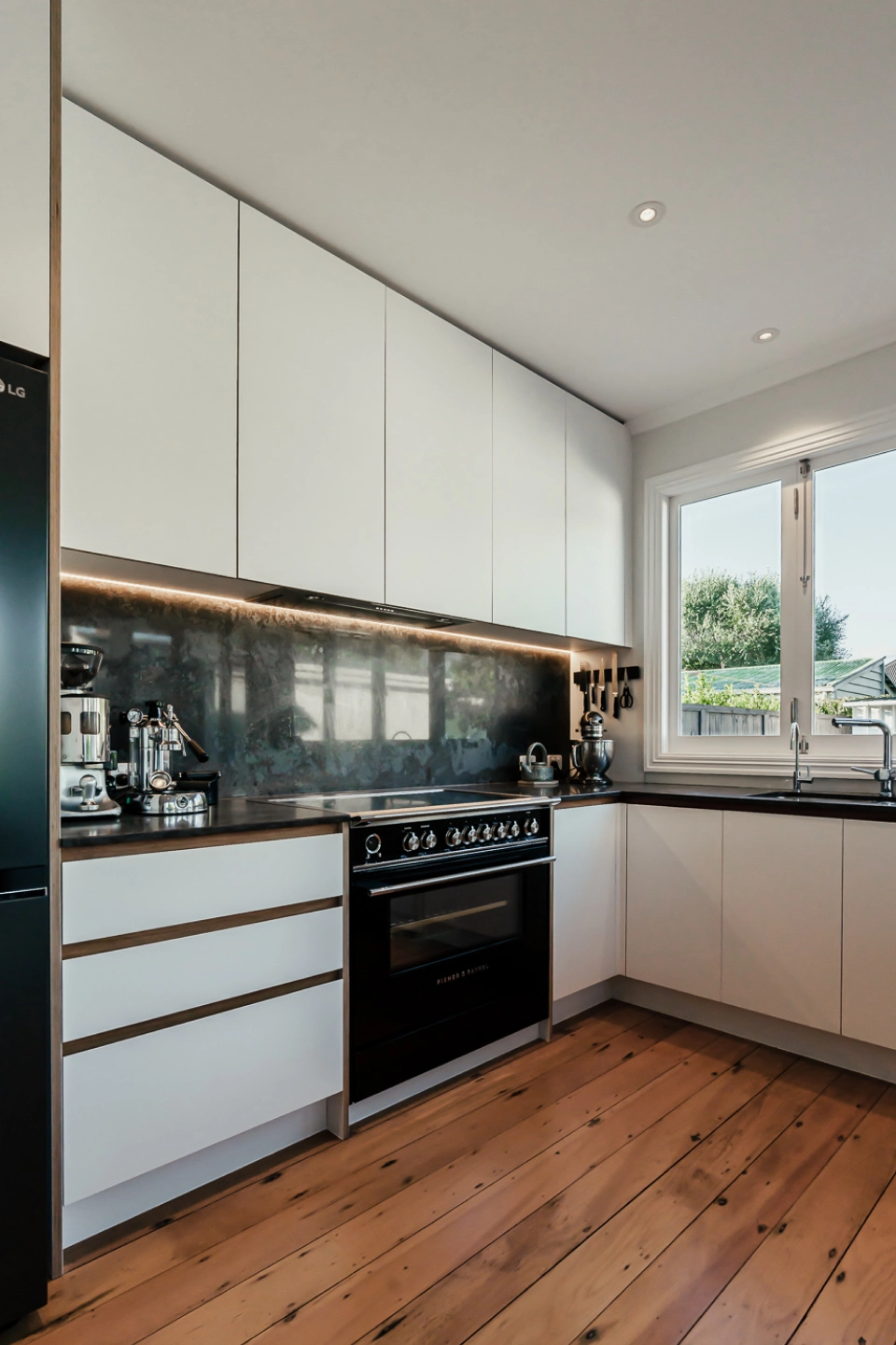 A sleek corner view of a modern kitchen with white cabinetry, black appliances, and warm wood flooring.