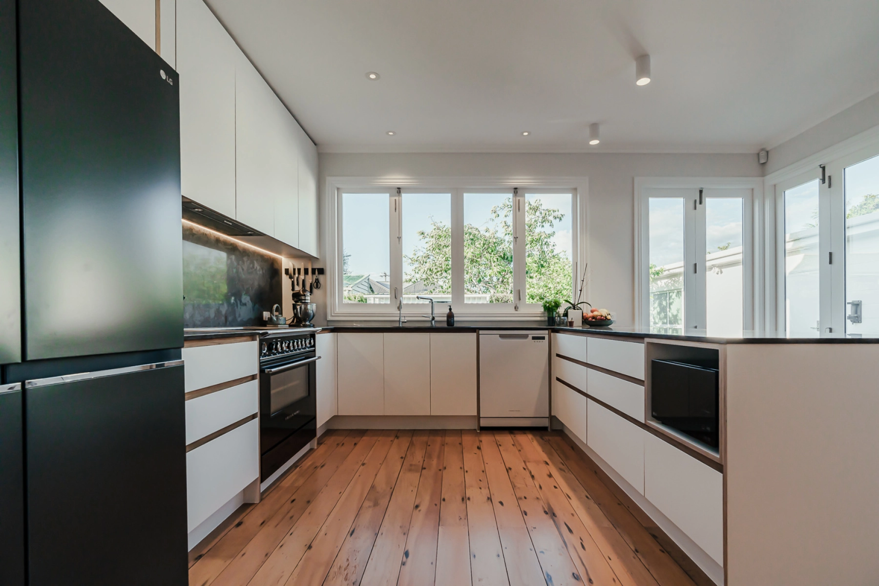 A bright, open kitchen with large windows, minimalist white cabinets, and black countertops.