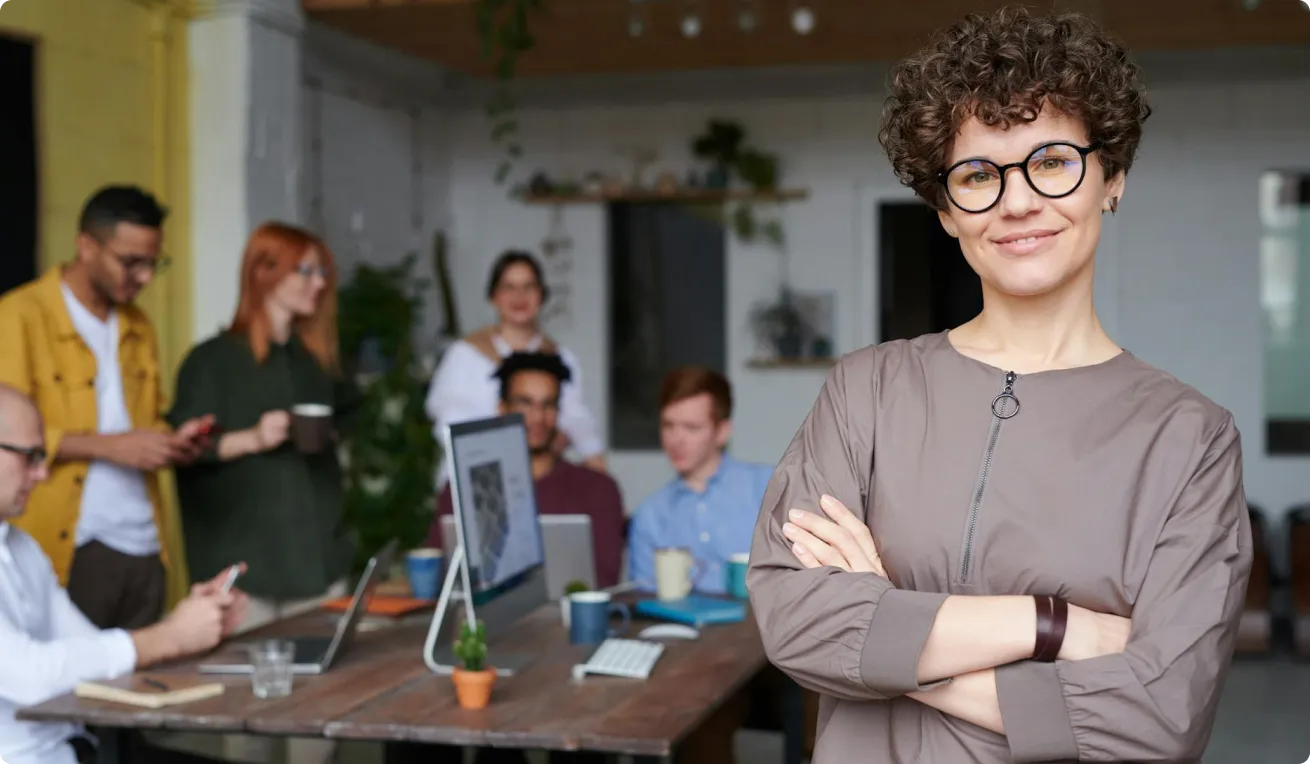 Woman with glasses and short curly hair is standing in front of people which are sitting and standing around a table
