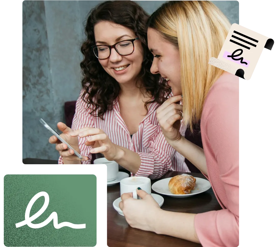 Two girls are sitting, smiling, looking at a mobile phone, eating a croissant and drinking coffee