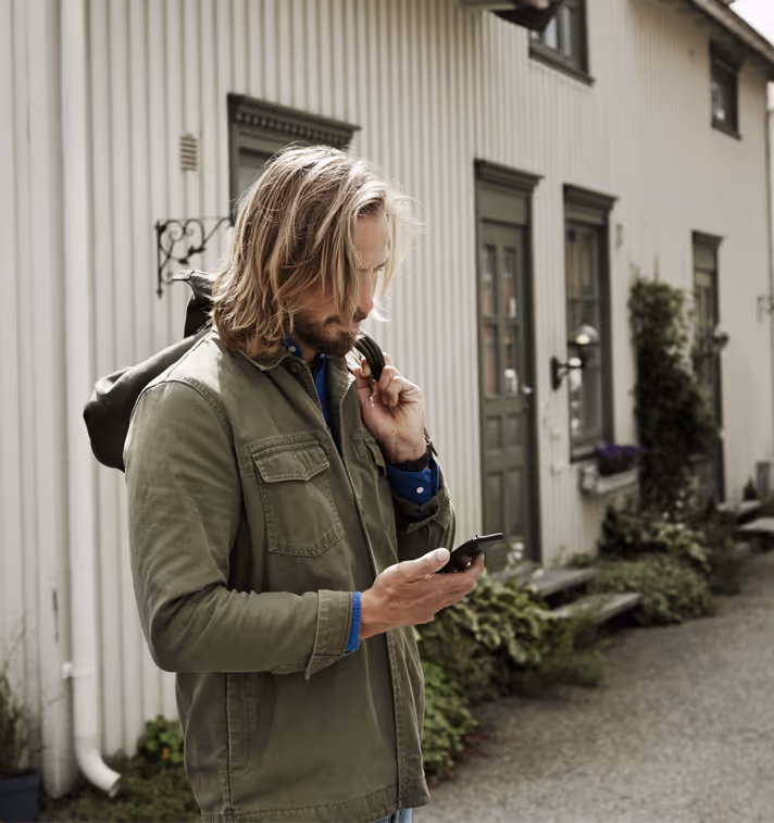 En middelaldrende mann i grønn jakke står og ser ned på en smarttelefon, i en stille gate som utstråler en rolig atmosfære.