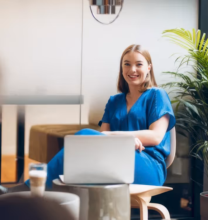 En smilende ung kvinne i blå kjole sitter avslappet foran en laptop på et lavt bord, omgitt av moderne møbler og planteliv i et lyst og innbydende rom.