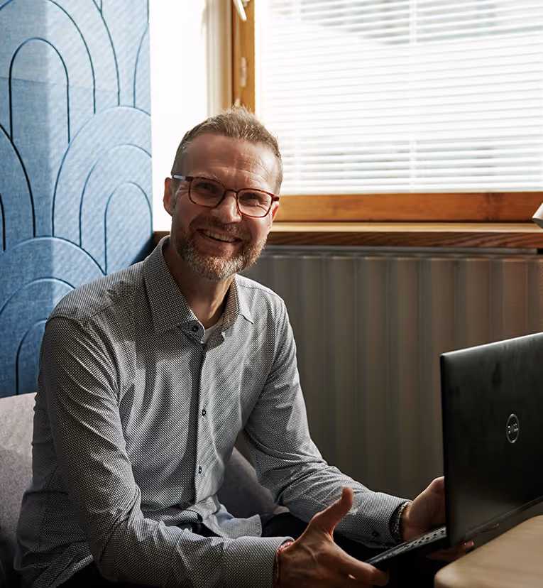 En middelaldrende mann smiler mot kameraet, sittende ved et skrivebord med en laptop.