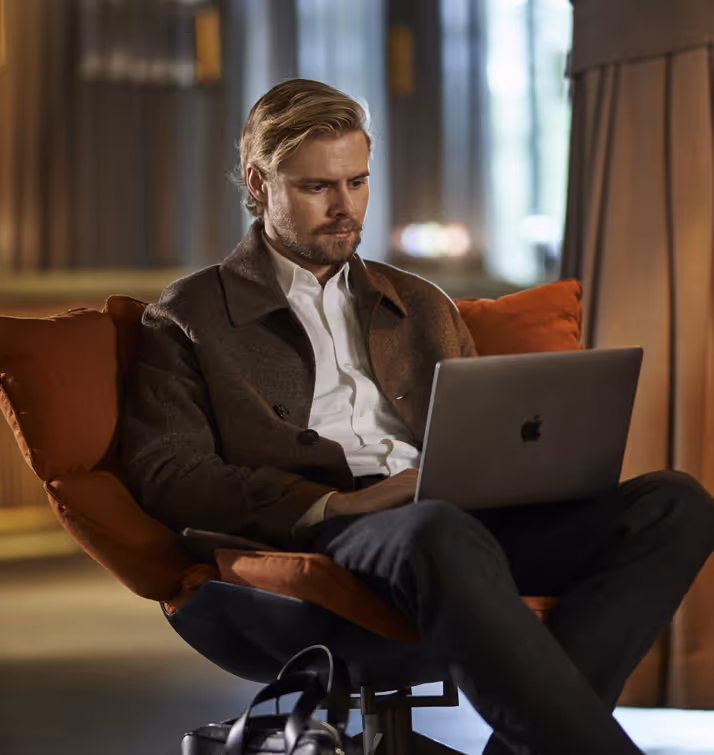 En velkledd mann sitter i en lounge-stol med en laptop i fanget og en konsentrert mine.