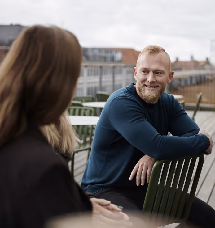 En mann sitter avslappet på en stol mens han diskuterer noe med en brunhåret kvinne vi kan se i forgrunnen.