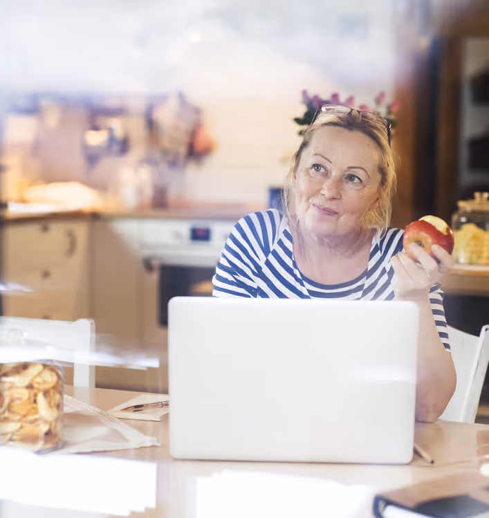 En middelaldret dame sitter i et lyst kontorlandskap med en åpen laptop foran seg mens hun ettertenksomt spiser et eple.