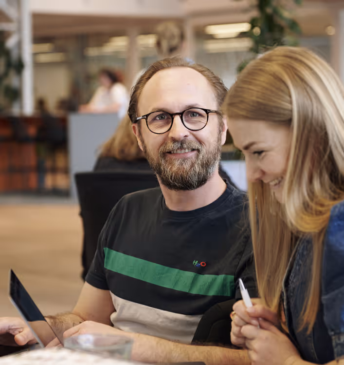 En middelaldret mann med briller ser mot kamera. Han og en blond kvinne sitter og går diskuterer noe på en laptop i et avslappet kontormiljø.