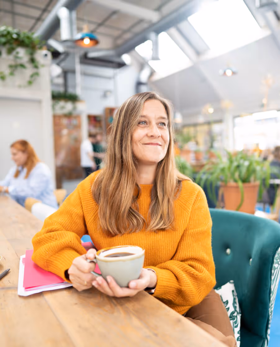 Woman in an orange sweater sat at table drinking a hot drink