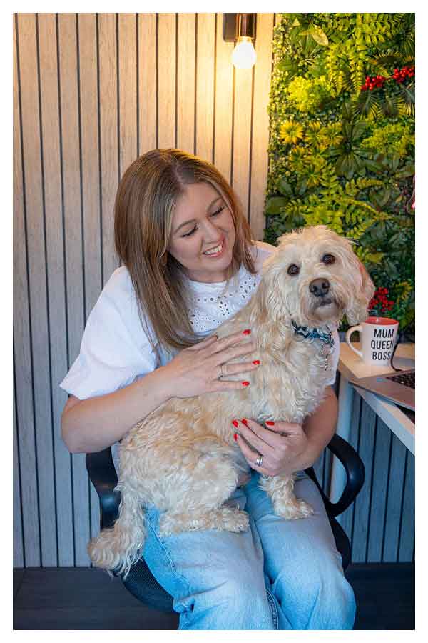 Kath, wedding celebrant, sitting in chair holding a beige curly-haired dog on her lap, with a green plant wall and mug reading 'MUM QUEEN BOSS' in the background.
