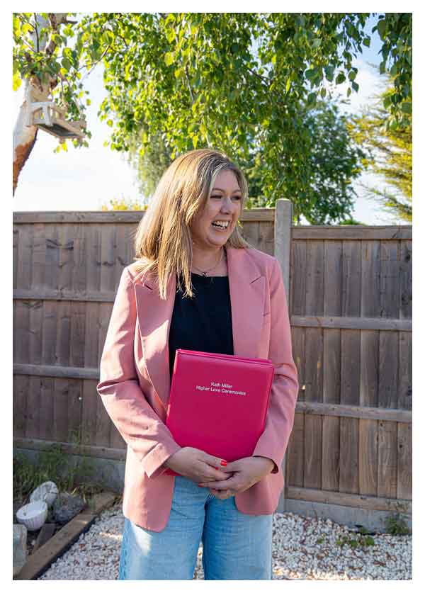 Kath, wedding celebrant in a pink blazer holding a red folder outdoors in front of a wooden fence with green trees overhead.