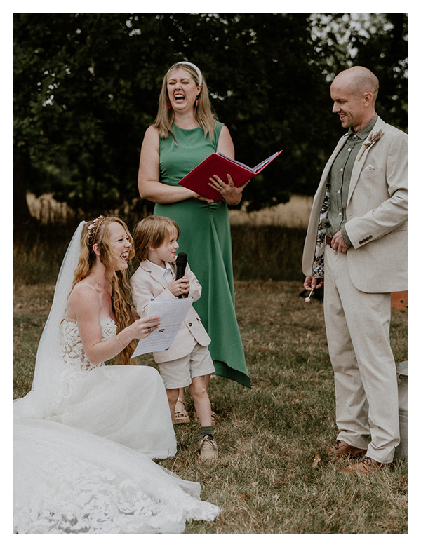 Bride kneeling on grass holding paper and smiling while a young boy with a microphone stands beside her, Kath, wedding celebrant in a green dress holding a red book behind them, and a man in a beige suit looking on.