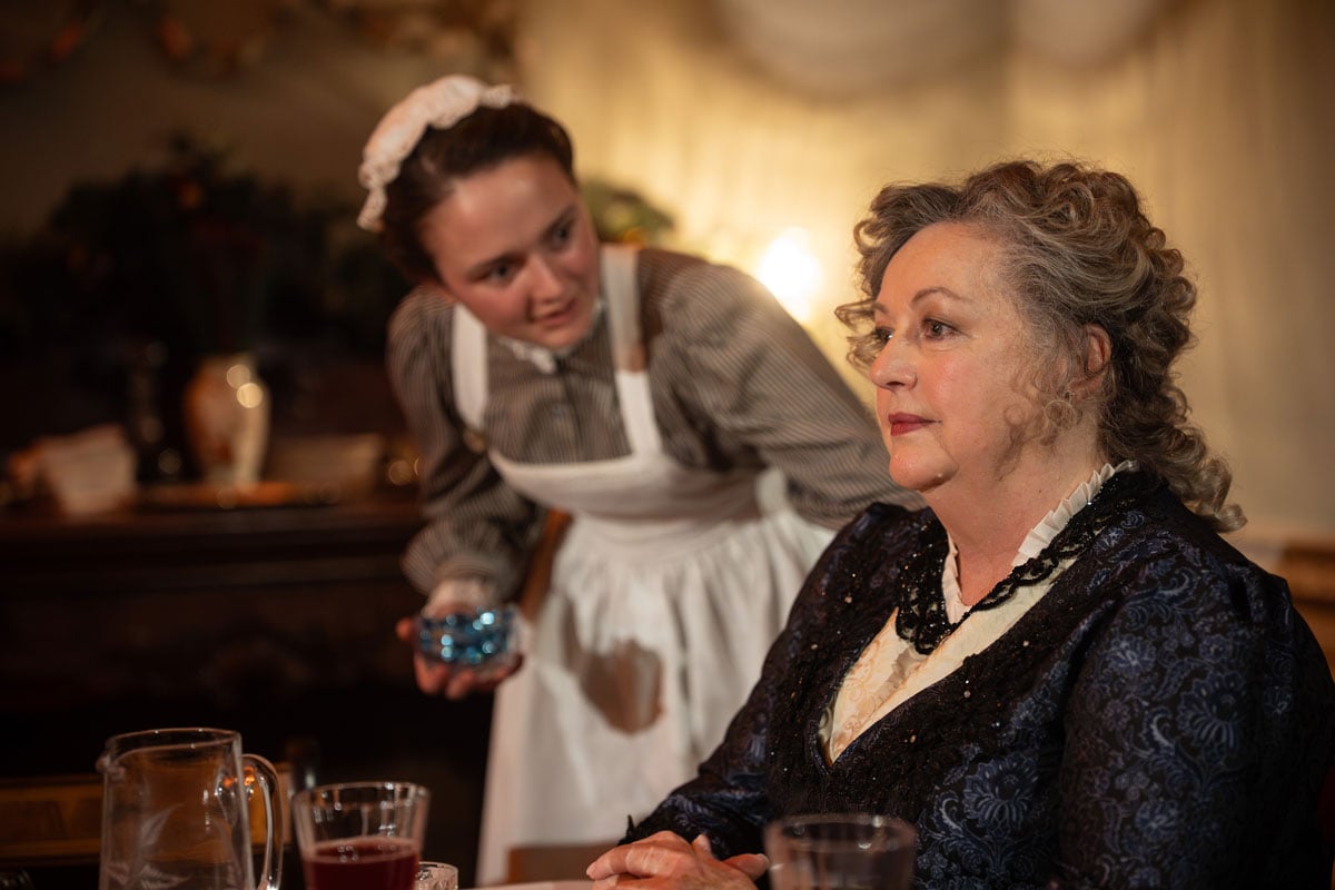 A serving girl leans in to enquire with an older woman