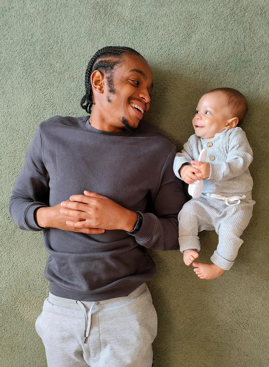 Man and baby lying on carpet facing each other, both smiling and making eye contact.