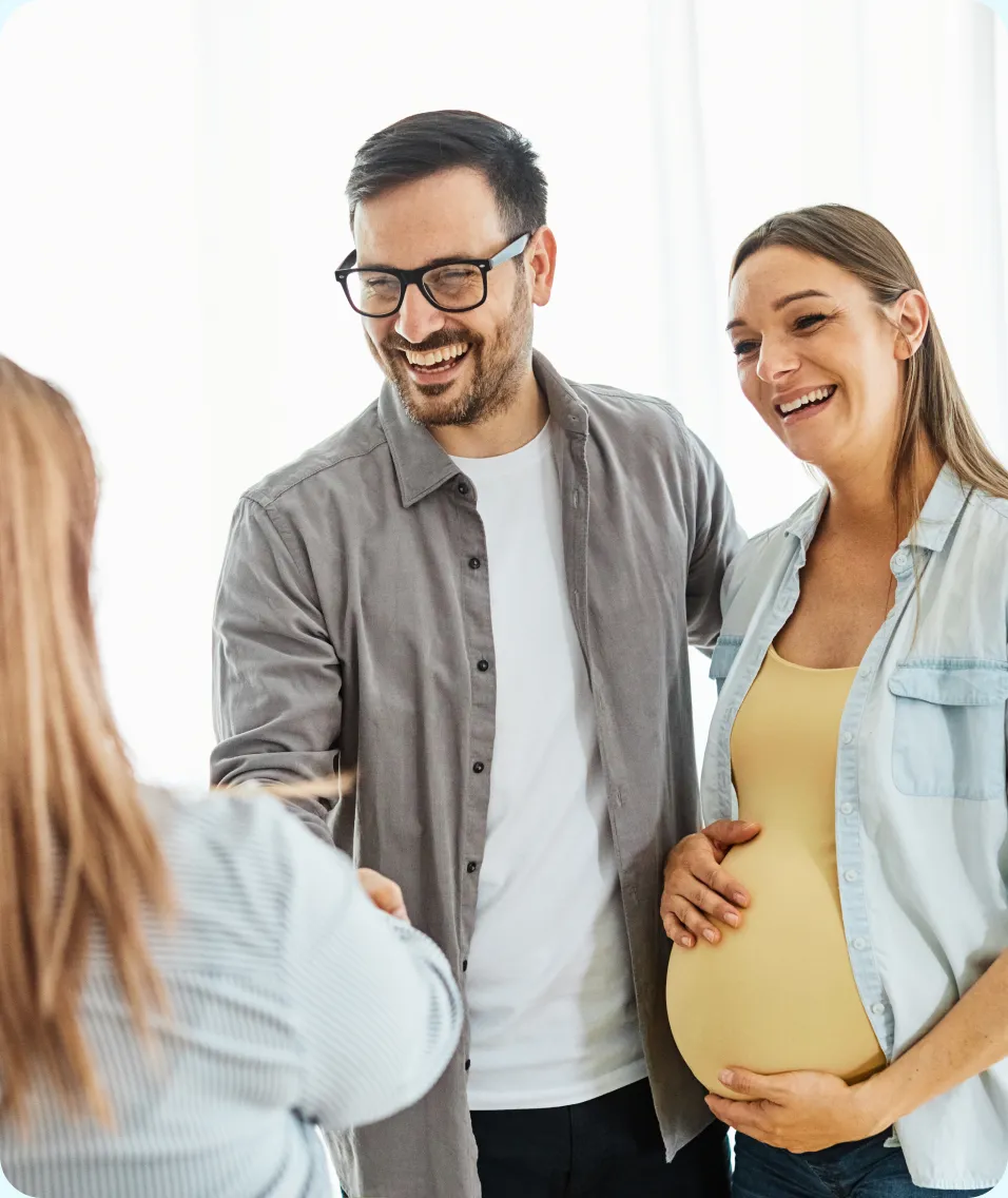 A smiling couple, with the woman visibly pregnant, greeting another person indoors.