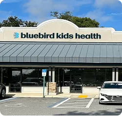Entrance of Bluebird Kids Health clinic with parking spaces and clear blue sky.