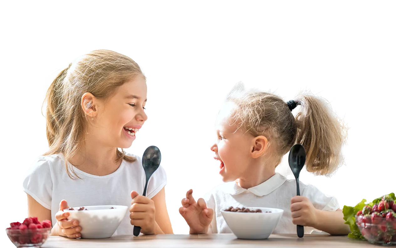 Two young girls smiling and holding spoons while sitting at a table with bowls of cereal and fruit.