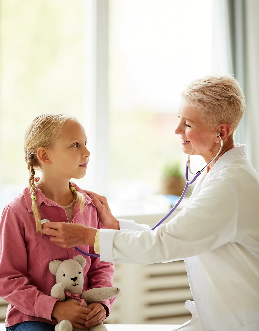 Smiling female doctor using a stethoscope to examine a young girl holding a teddy bear in a bright medical office.
