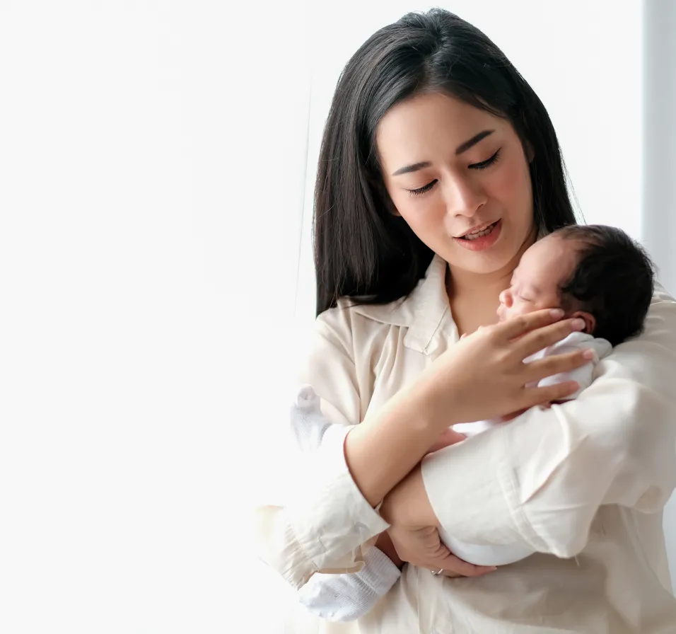 Mother gently cradling and looking down at her sleeping newborn baby wrapped in a white outfit.