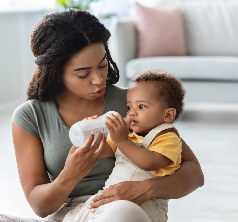 Mother bottle-feeding a baby boy sitting on her lap in a bright living room.