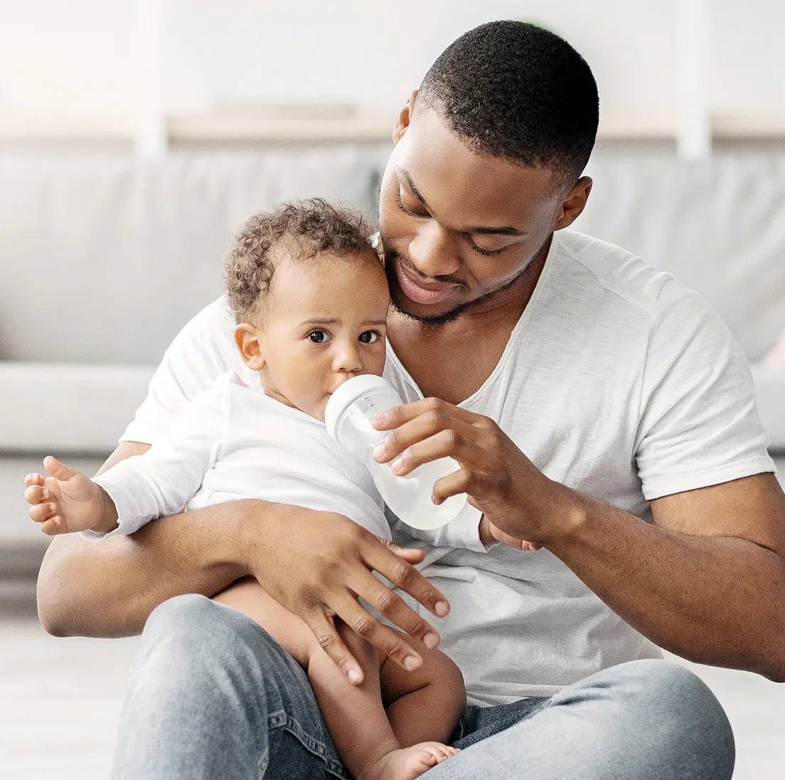 Father in a white shirt feeding baby with a bottle while sitting on the floor.
